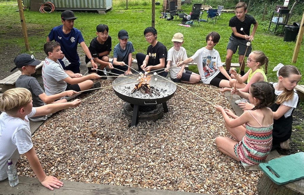 Group of children gathered around a campfire at a party, happily cooking food on sticks, with smiling faces and trees in the background.