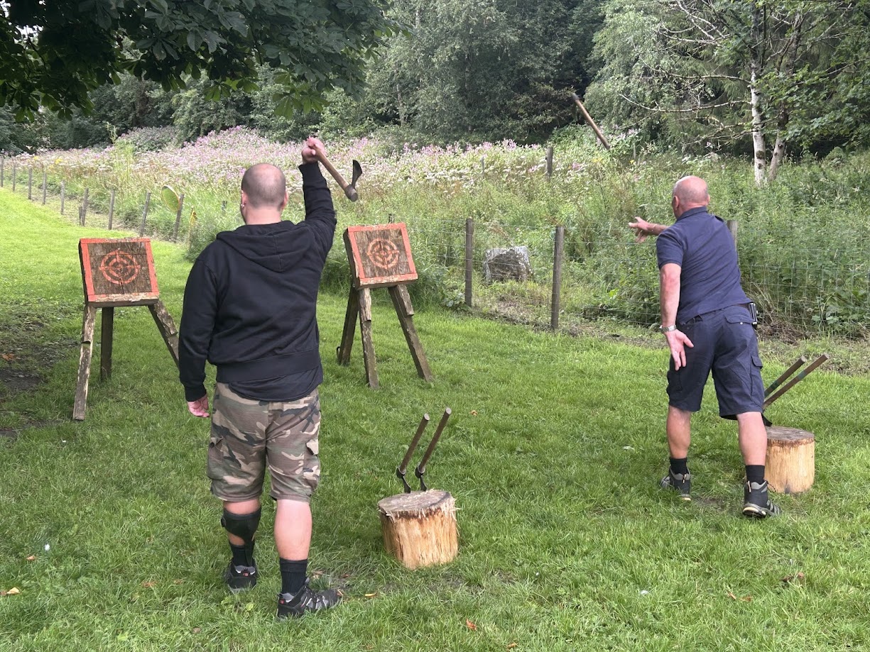Two adults engaged in tomahawk throwing at Wood-Sage Outdoor, each aiming at wooden target boards set on grassy terrain. One person is mid-throw while the other prepares to release a tomahawk, with spare axes resting on tree stumps nearby