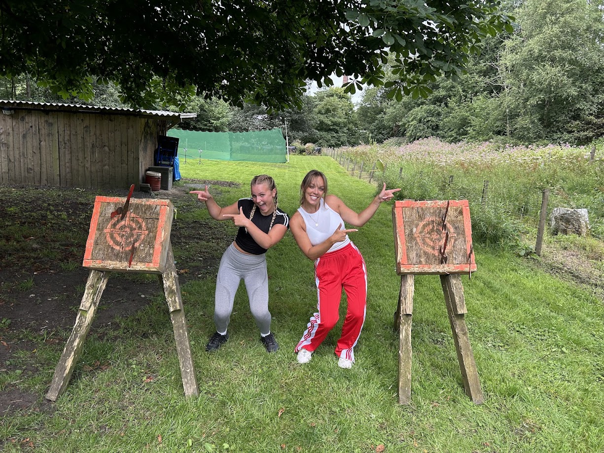 Two smiling participants posing on our grassy tomahawk range at Wood-Sage Outdoor, each pointing proudly at their target