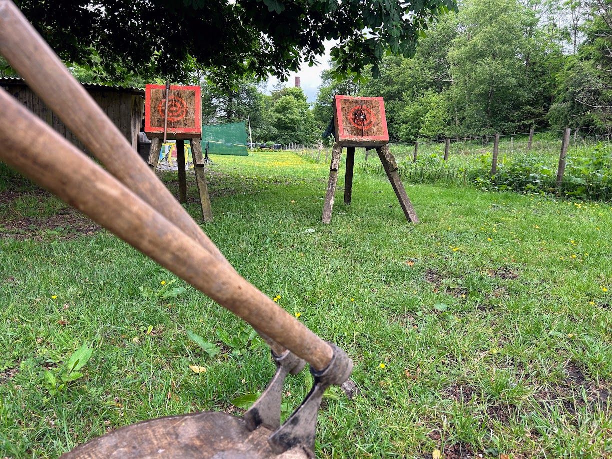 Outdoor tomahawk throwing setup at Wood-Sage Outdoor. In the foreground, there is a wooden stump with two tomahawks embedded in it. In the background, there are two wooden targets on stands. 