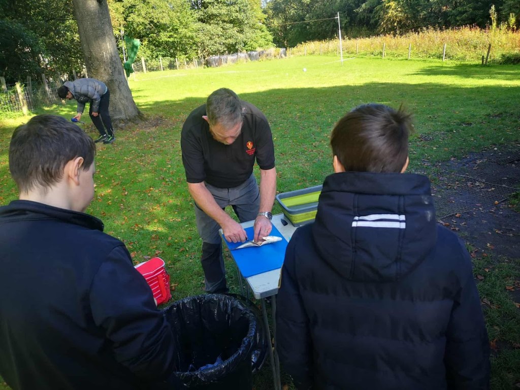 earning outdoor cooking skills by preparing a whole fish over a campfire, seasoning it carefully before cooking on a stick, surrounded by nature.