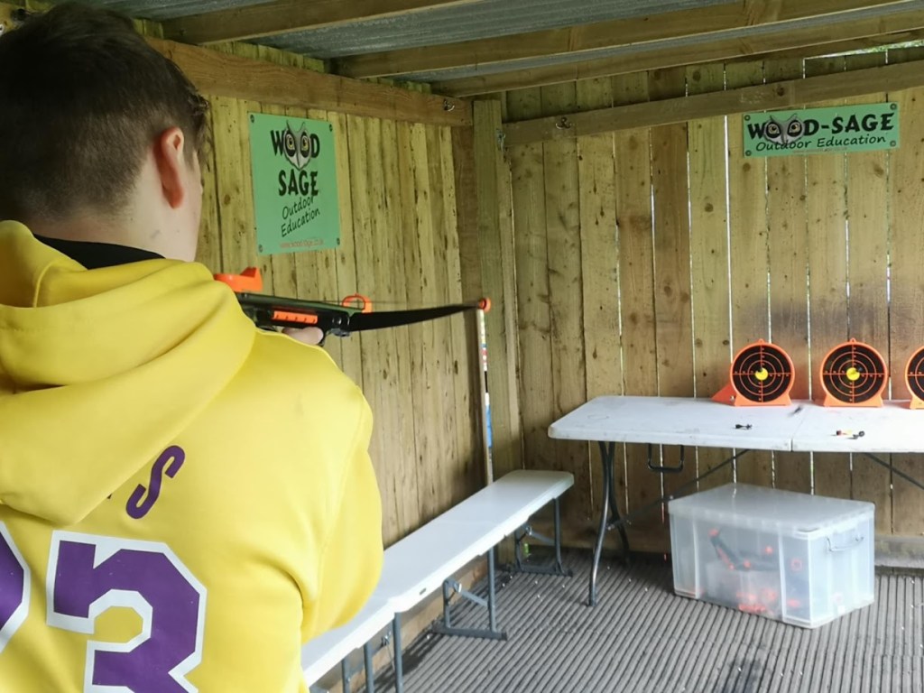 A young person aiming a soft-tipped crossbow at a target outdoors, guided by an instructor, practicing focus and coordination in a safe, fun activity
