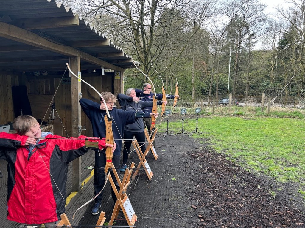 Four people practising archery under a wooden shelter.