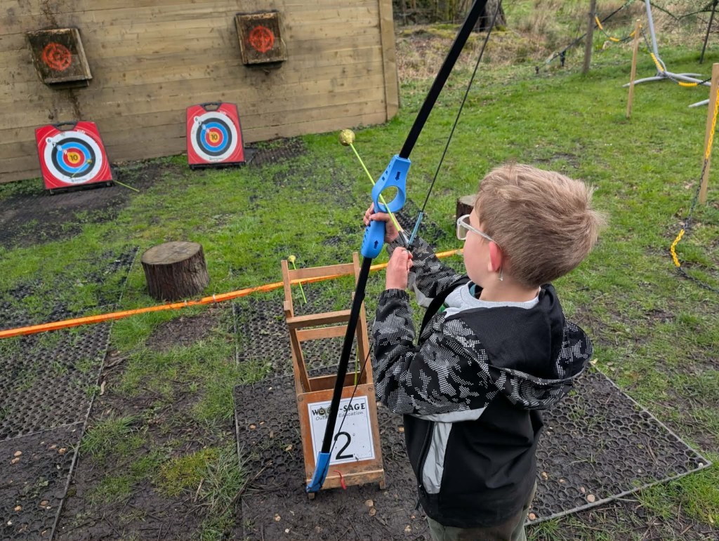 Child taking aim with a bow during Easter HAF 2026 at Wood‑Sage Outdoor Education, with colourful archery targets in the background.
